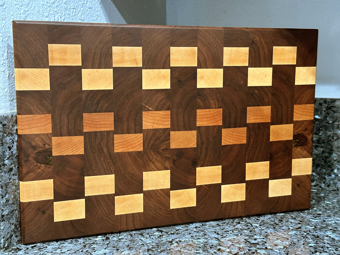 Crafted from the finest hardwoods in North America, this end grain cutting board features rich black walnut with cherry and maple accents. Chamfered edges and finger grooves give a modern twist to the classic cutting board styling. Hard rubber feet gives the chef a stable worksurface along with providing air circulation under the board, allowing it to dry after washing. Board dimensions: 18"L x 12"W x 0.5"H.