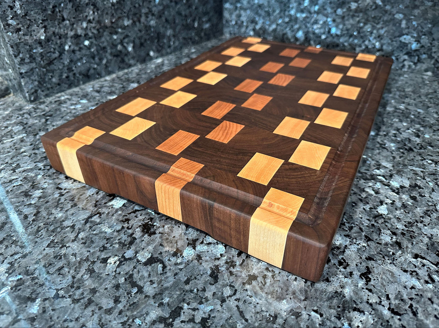 Crafted from the finest hardwoods in North America, this end grain butcher block features rich black walnut with cherry and maple accents. Chamfered edges and finger grooves give a modern twist to the classic butcher block styling. Hard rubber feet gives the chef a stable worksurface along with providing air circulation under the block, allowing it to dry after washing. Board dimensions: 18"L x 12"W x 1.75"H. As a handcrafted product, please allow for approximately 1/4" variation in dimensions.