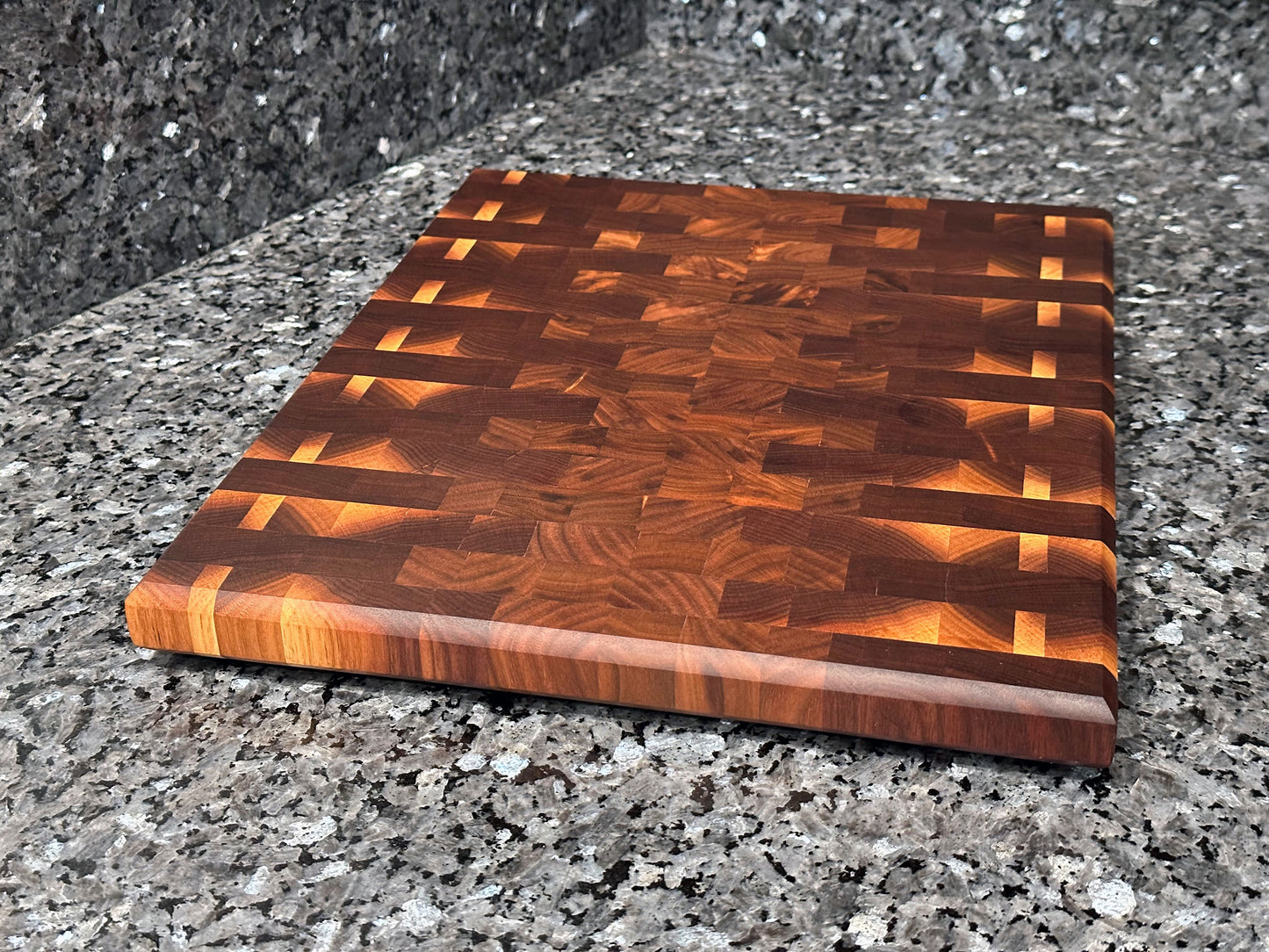 Crafted from the finest hardwoods in North America, this mosaic inspired end grain cutting board features rich black walnut. Chamfered edges give a modern twist to the classic cutting board styling. Hard rubber feet gives the chef a stable worksurface along with providing air circulation under the board, allowing it to dry after washing. Board dimensions: 14.75"L x 12"W x 1.25"H. As a handcrafted product, please allow for approximately 1/4" variation in dimensions.