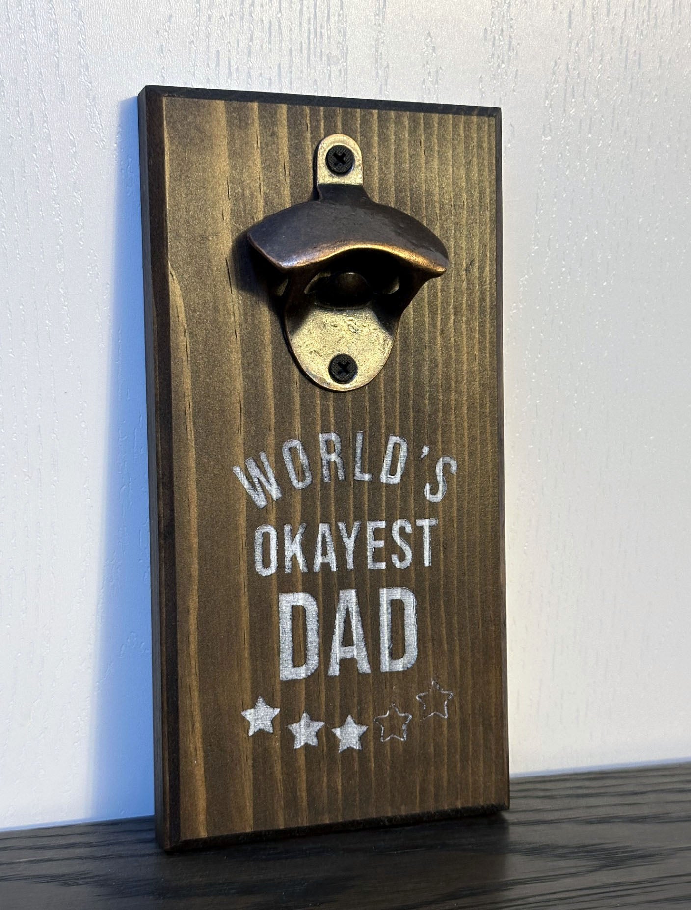 Custom magnetic bottle opener featuring a "World's Okayest Dad" theme. Each bottle opener is handmade and has hidden rare earth magnets to catch metal bottle caps. Wall-mounted or magnetic backing options. Made by Space Rubber Ducky Designs in Southern California, USA.