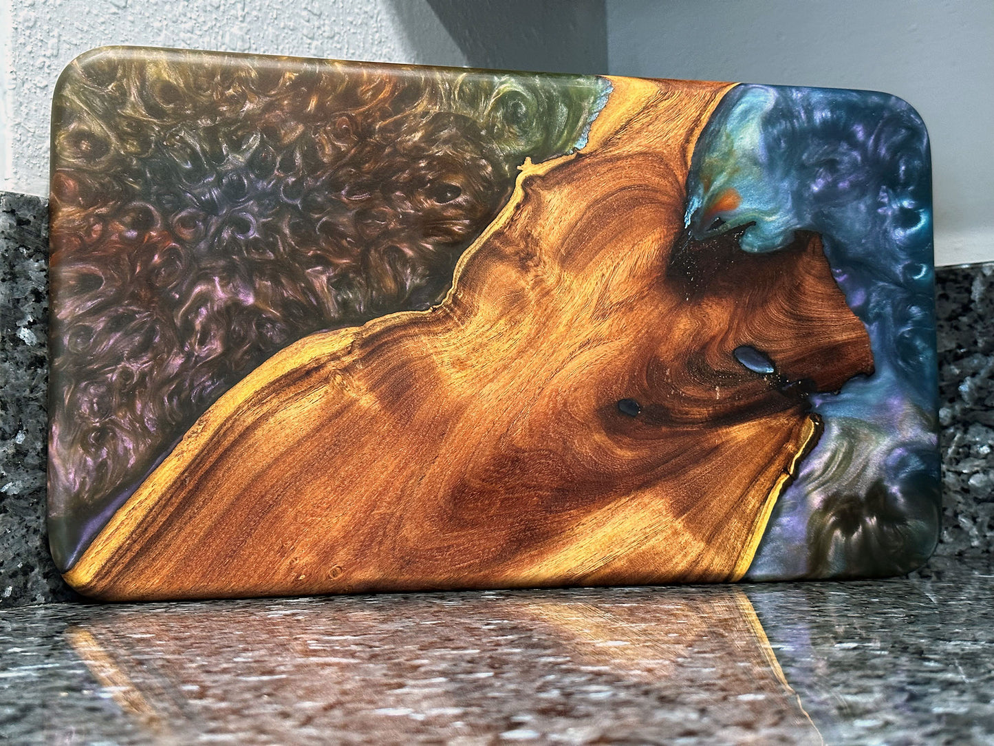 The natural beauty of this live edge mesquite serving board is accented with a blast of color. Dark blues and purples swirl together like a galactic nebula while a bubbling sea of color hypnotizes. Food safe epoxy resin was used to create this design. Using knives or other sharp utensils on the epoxy resin surface may damage it and is not recommended. Board dimensions: 17"L x 9.75"W x 0.75"H. As a handcrafted product, please allow for approximately 1/4" variation in dimensions.