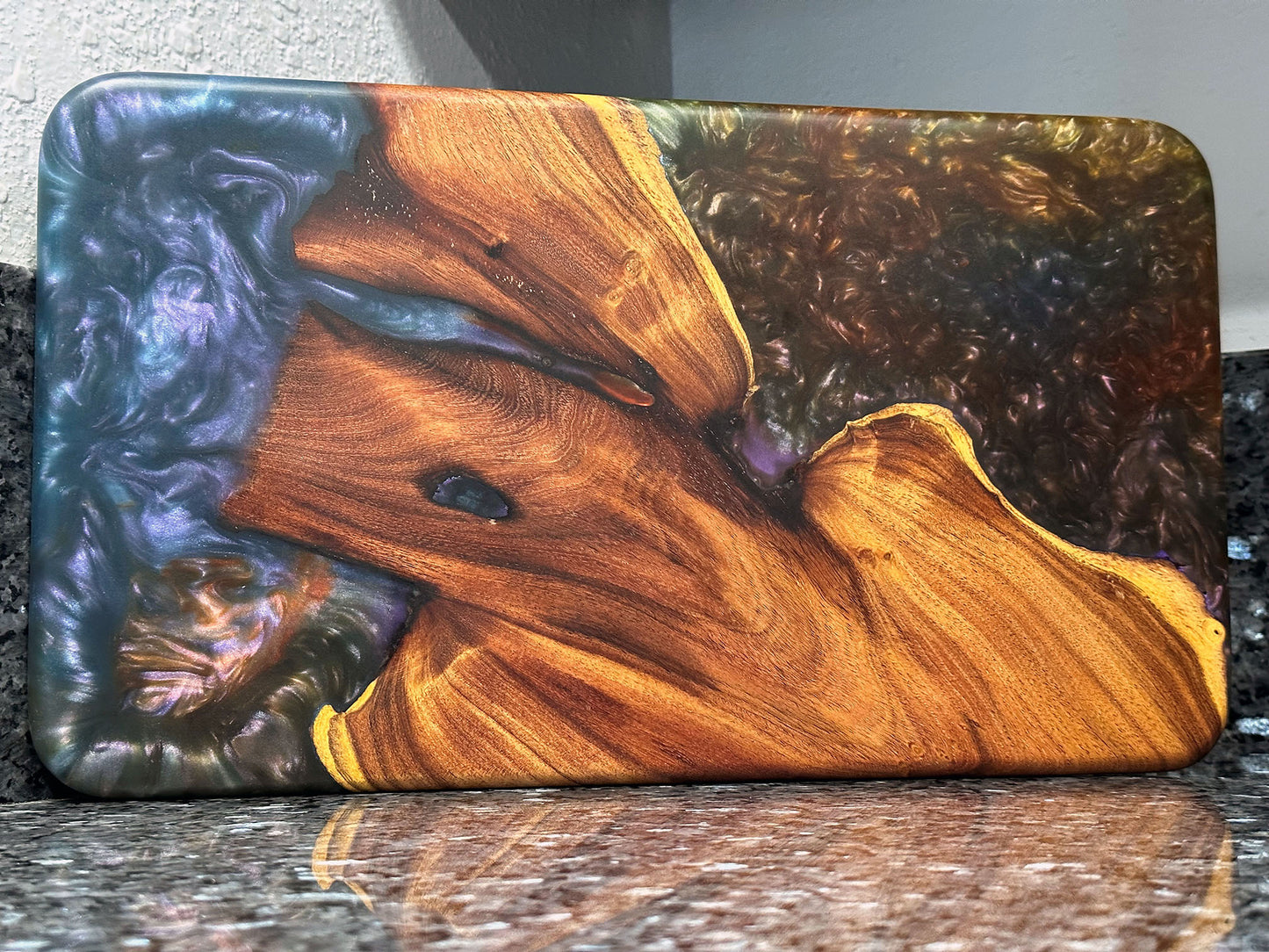 The natural beauty of this live edge mesquite serving board is accented with a blast of color. Dark blues and purples swirl together like a galactic nebula while a bubbling sea of color hypnotizes. Food safe epoxy resin was used to create this design. Using knives or other sharp utensils on the epoxy resin surface may damage it and is not recommended. Board dimensions: 17"L x 9.75"W x 0.75"H. As a handcrafted product, please allow for approximately 1/4" variation in dimensions.