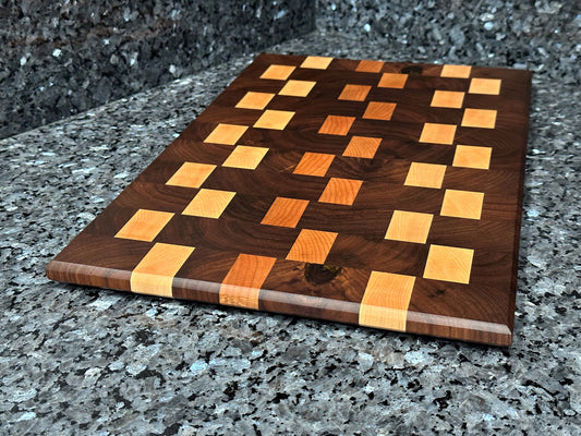 Crafted from the finest hardwoods in North America, this end grain cutting board features rich black walnut with cherry and maple accents.  Chamfered edges and finger grooves give a modern twist to the classic cutting board styling. Hard rubber feet gives the chef a stable worksurface along with providing air circulation under the board, allowing it to dry after washing.  Board dimensions:  18"L x 12"W x 0.5"H.