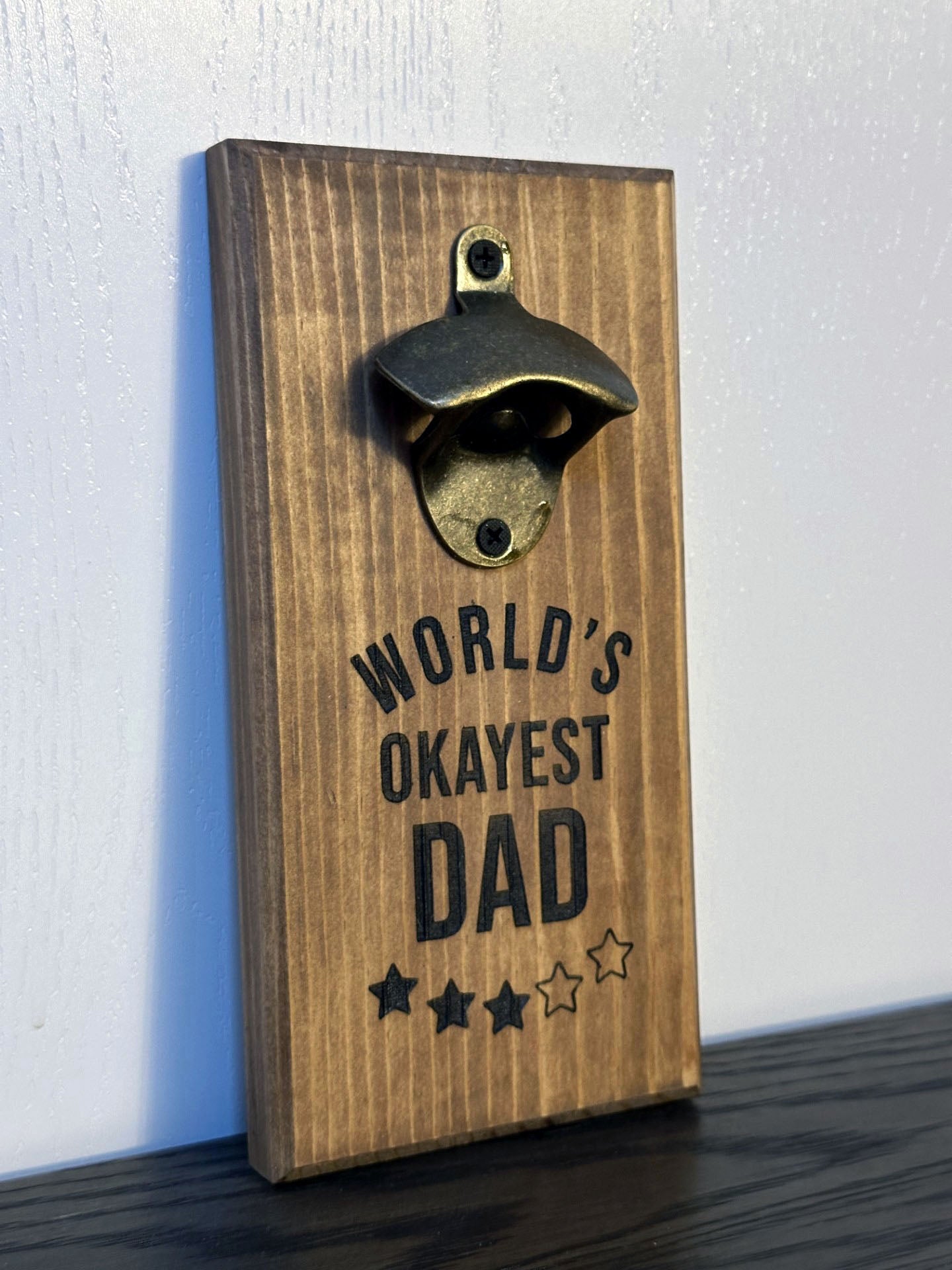 Custom magnetic bottle opener featuring a "World's Okayest Dad" theme. Each bottle opener is handmade and has hidden rare earth magnets to catch metal bottle caps. Wall-mounted or magnetic backing options. Made by Space Rubber Ducky Designs in Southern California, USA.